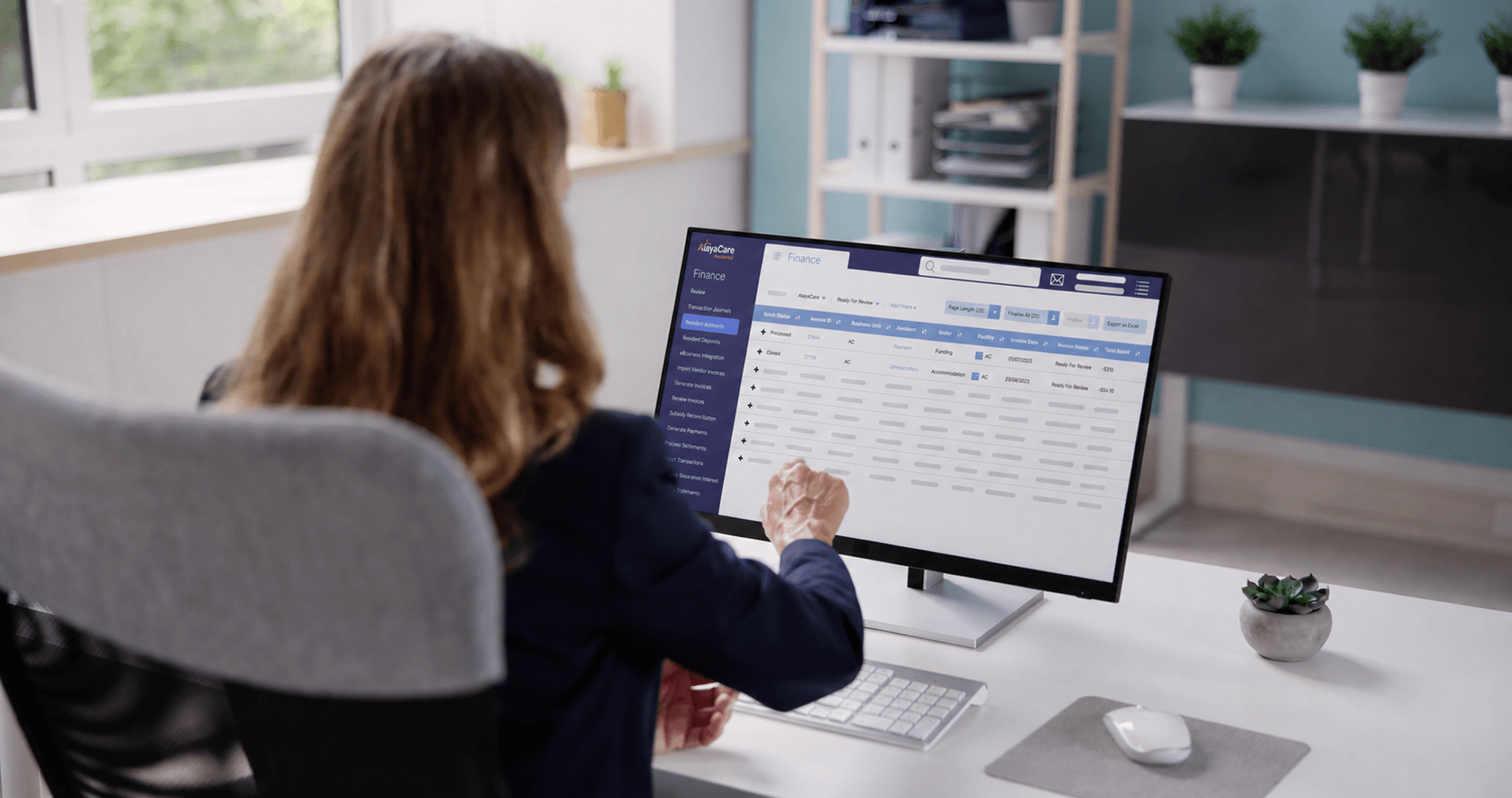 Office worker viewing an AlayaCare Finance dashboard showing a resident invoice batch table with review status and totals